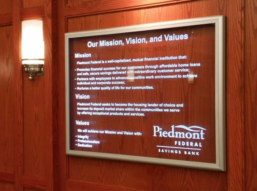 A framed glass display on a wooden wall showing Piedmont Federal's mission, vision, and values. The display has carved text and logo on it.