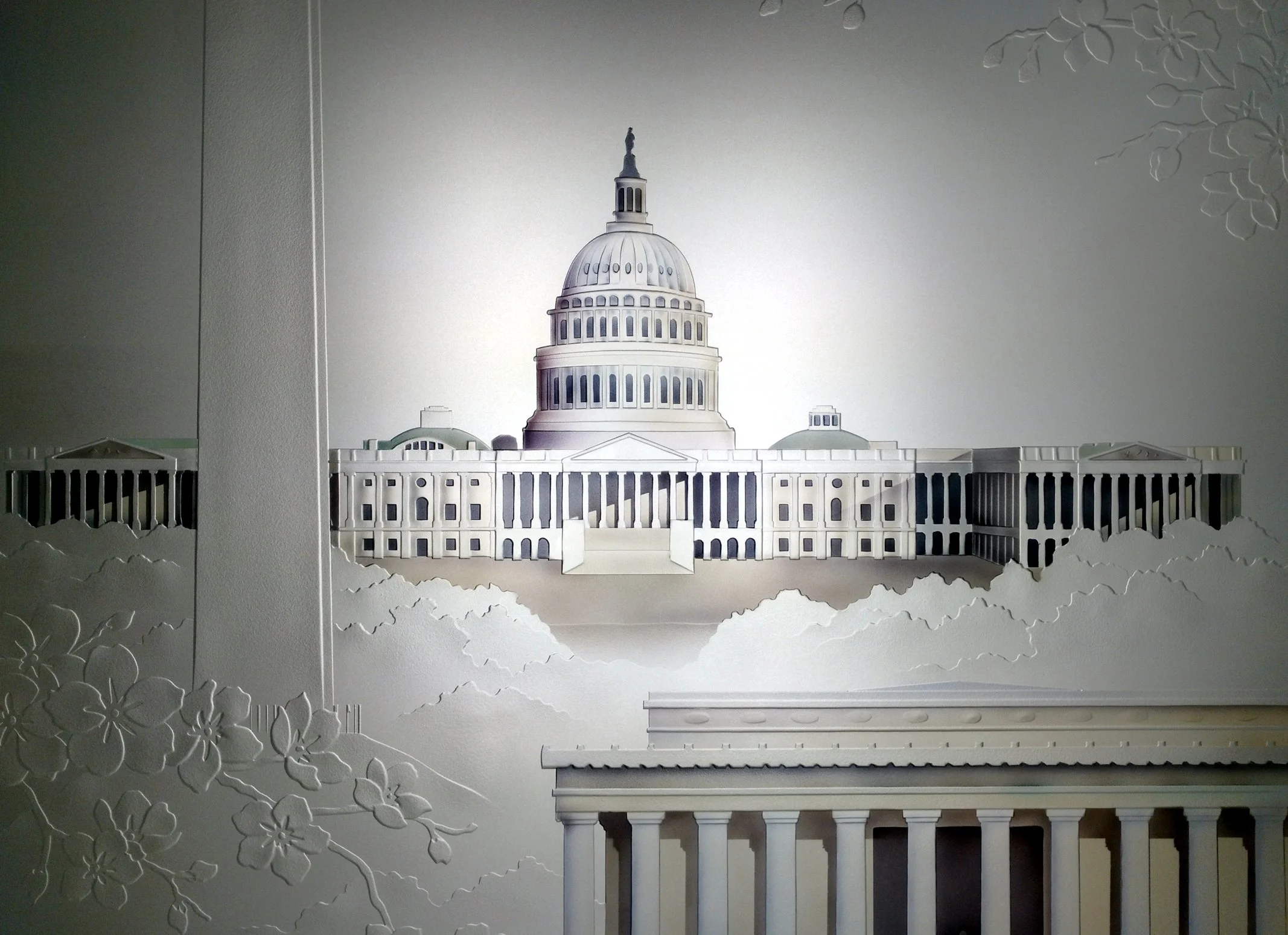 White layered paper sculpture of the U.S. Capitol building with floral embossing on the wall behind it.
