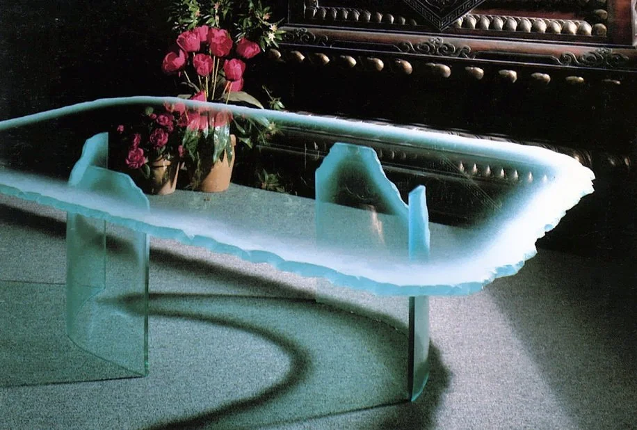 A glass cocktail table with a frosted, uneven edge supported by two curved glass bases, with a potted plant of pink flowers in the background and ornate dark wood furniture.