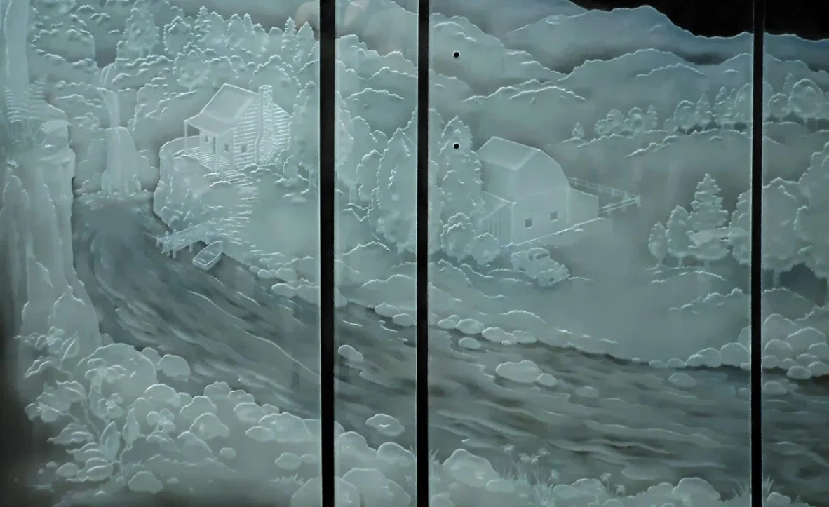 Carved shower door glass art depicting a rural landscape with trees, a river, and a fishing cabin.