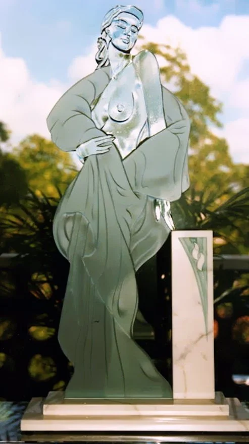 Glass sculpture of a woman, draped in fabric, standing outdoors with trees and a blue sky in the background.