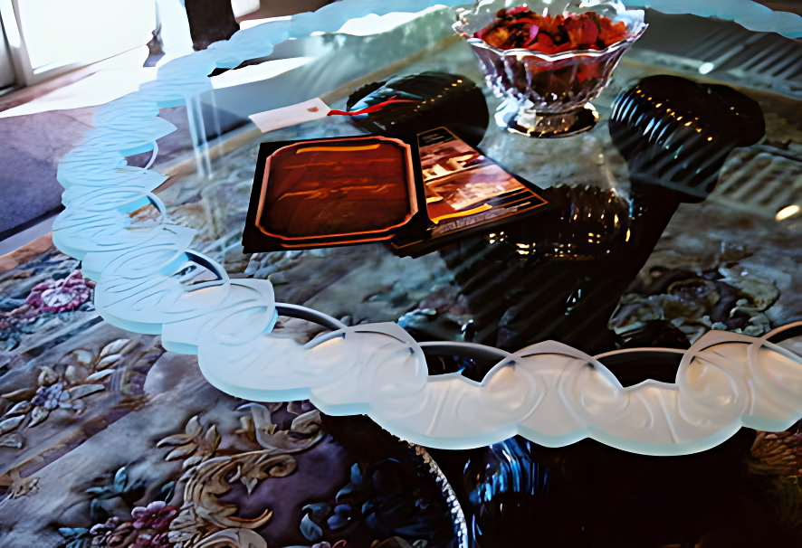A glass table with a white ruffled edge, holding a bowl of strawberries, a couple of rectangular objects, and a small decorative black item, with a patterned cloth underneath.
