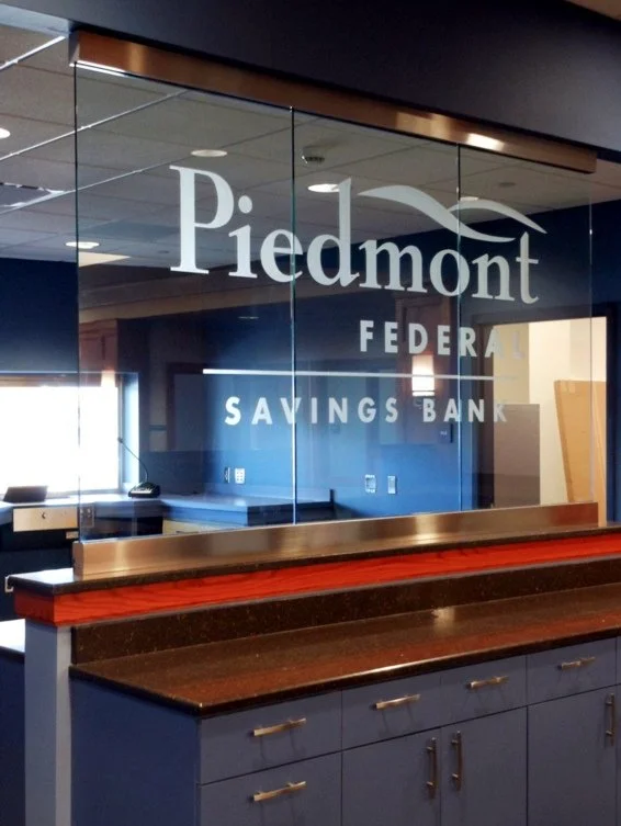 Interior of a bank with a glass wall bearing the logo and name 'Piedmont Federal Savings Bank' in sandblasted letters.