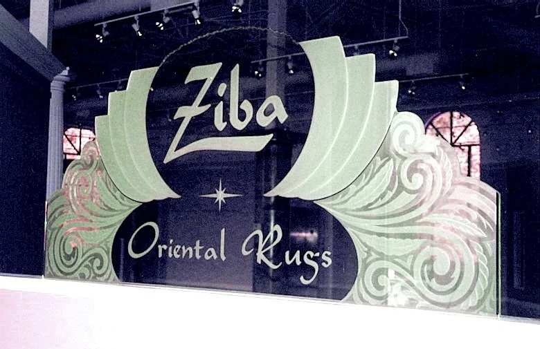 Shaped glass sign reading 'Ziba Oriental Rugs' with decorative patterns and swirls surrounding the text, inside a building with dark walls and high windows.