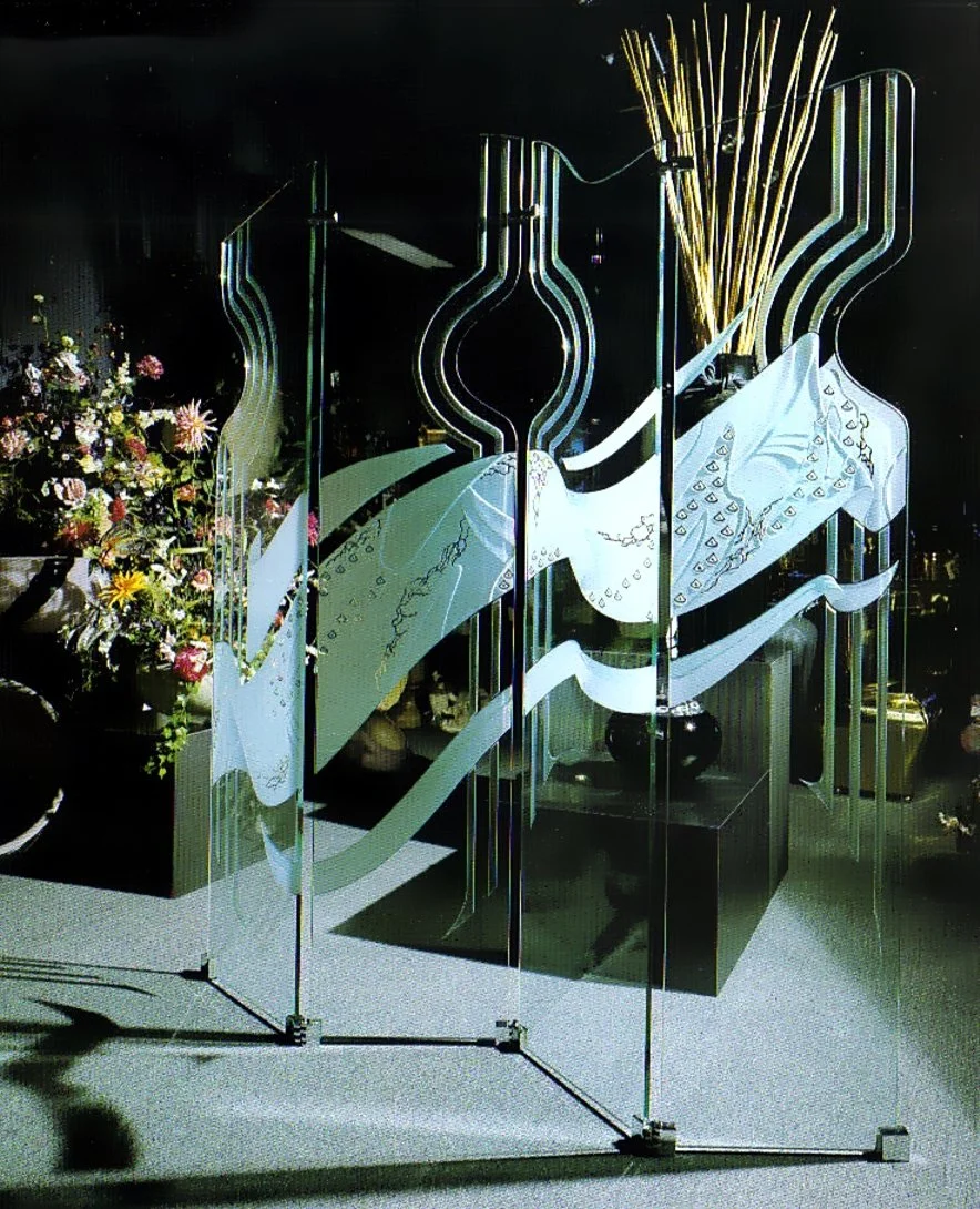 Decorative carved glass divider panels with abstract design, flanked by flower arrangements and a black pedestal, illuminated in a dark setting.