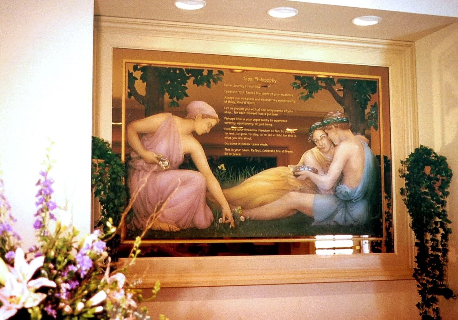 Glass painting of three classical mythological figures, two women and a man, surrounded by greenery and flowers, with a text panel about spa philosophy in the background.
