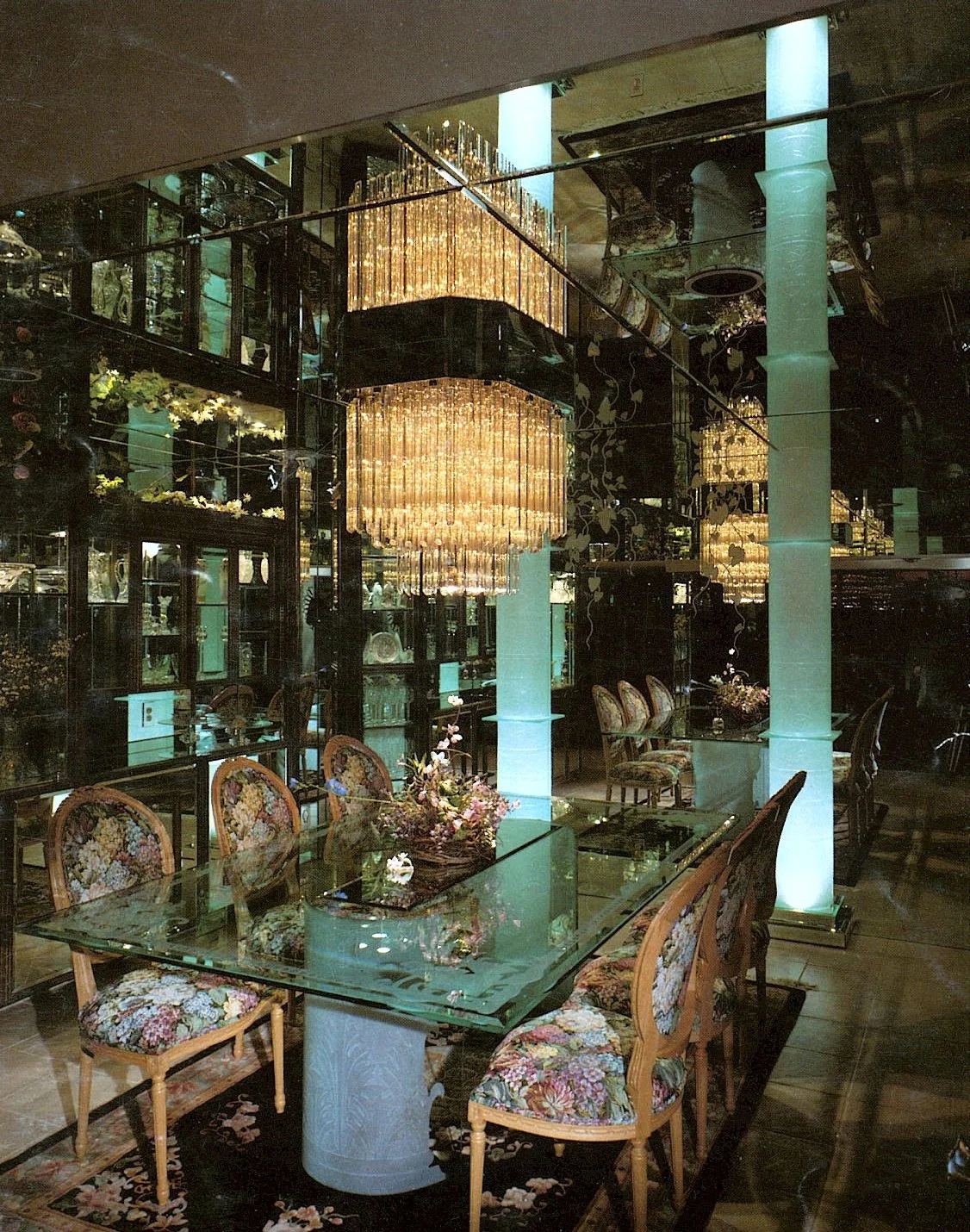Luxurious dining room with custom carved glass tabletop and bases, bent carved glass columns with back lighting, floral upholstered chairs, elegant floral centerpiece, large chandelier, and wall mirrors.