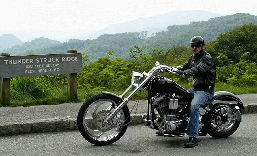 Man riding a black motorcycle on the Blue Ridge Parkway, near a sign that reads 'Thunder Struck Ridge, elevation 4780 feet, Green trees and hills in the background.