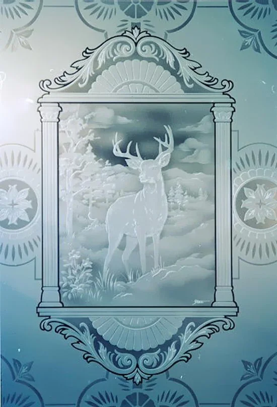 Etched and carved glass with a deer, with large antlers standing in a forest with trees, framed by ornate decorative borders.