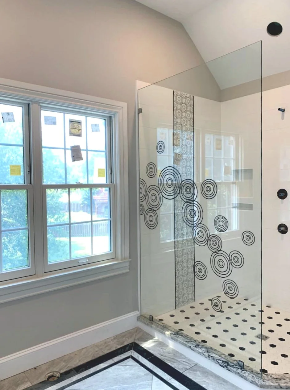 Bathroom shower area with a glass door decorated with black concentric circle patterns. Two windows with white framing are visible, showing trees outside. The bathroom features a light-colored wall, tiled floor with a border, and a small round drain on the floor.