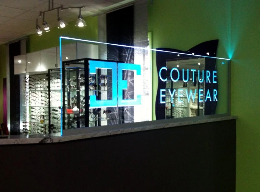 Store display with edge lit glass sign reading 'COUTURE EYEWEAR' next to a mirrored surface and a glass showcase with eyeglasses inside, set against green walls.