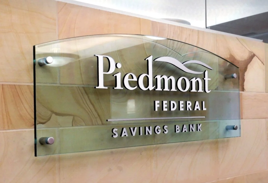 A deep carved and painted glass sign with the words 'Piedmont Federal Savings Bank' mounted on a beige marble wall.