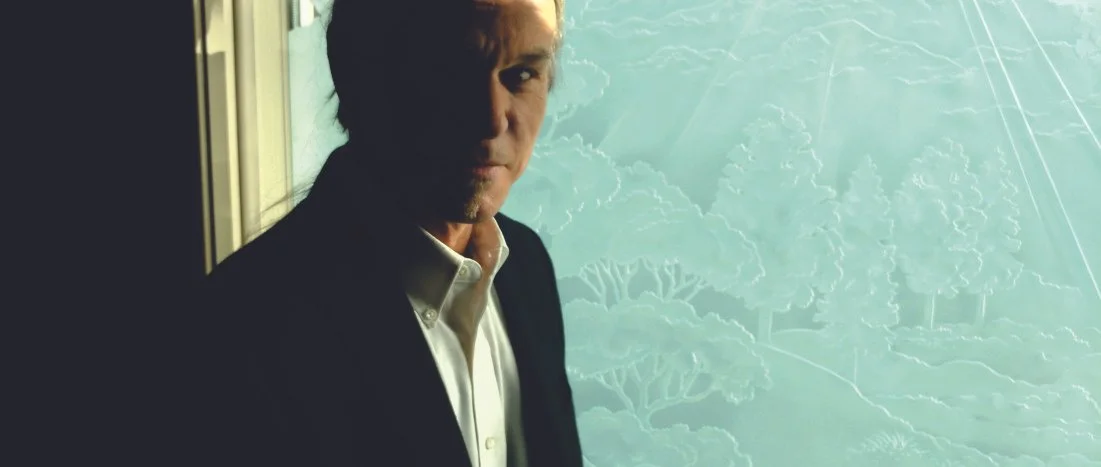 Man in a dark suit looking out a window with a light blue background featuring trees and rays of sunlight.