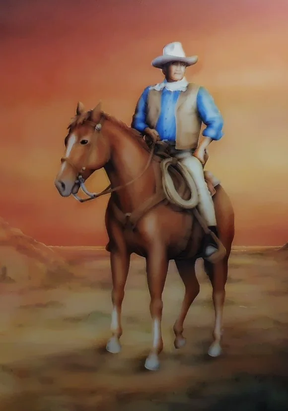 A carved glass painting of a cowboy wearing a wide-brimmed hat, blue shirt, vest, and chaps, riding a brown horse against a desert landscape with a warm, sunset sky.