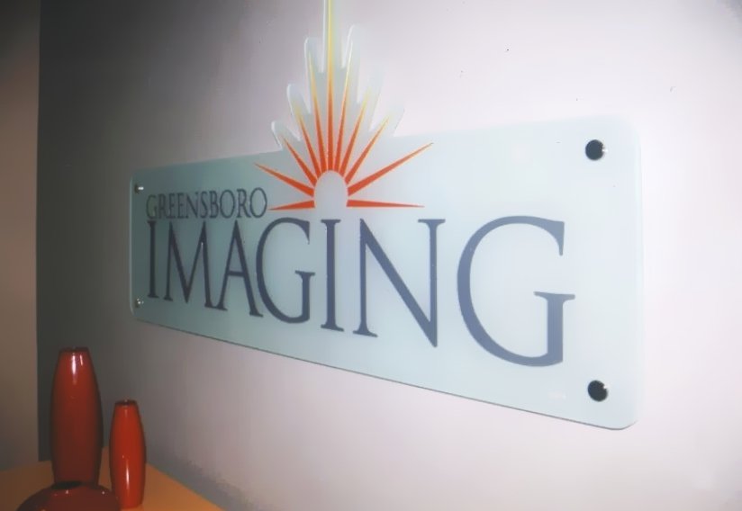 Sculpted glass sign reading 'Greensboro Imaging' with a sunrise design and three red vases on a table.