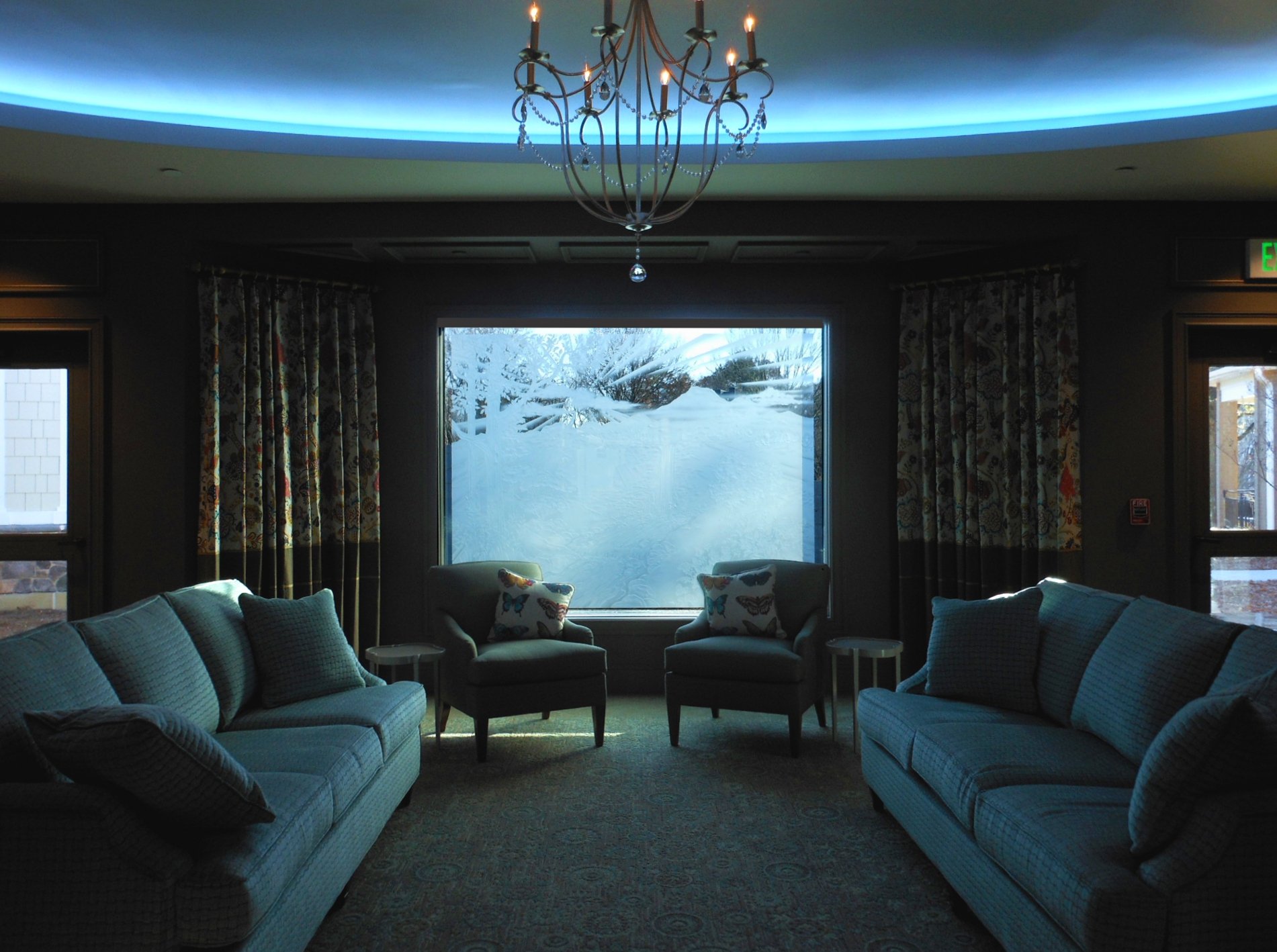 Indoor lounge with dark walls, two sofas, two armchairs with butterfly pillows, curtains, chandelier, large carved glass window.