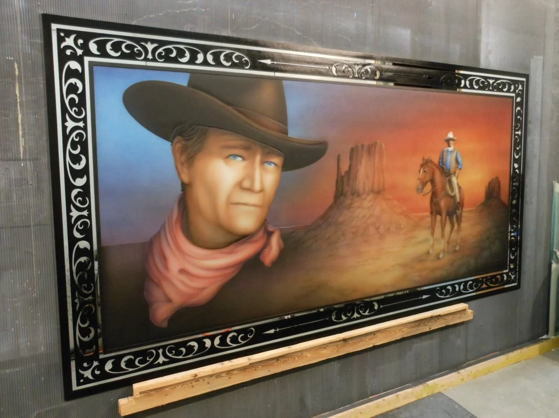 A painted Western scene featuring John Wayne with blue eyes wearing a large cowboy hat and a reddish neckerchief, with a desert landscape in the background including a prominent butte and a cowboy wearing a blue shirt and hat while riding a horse
