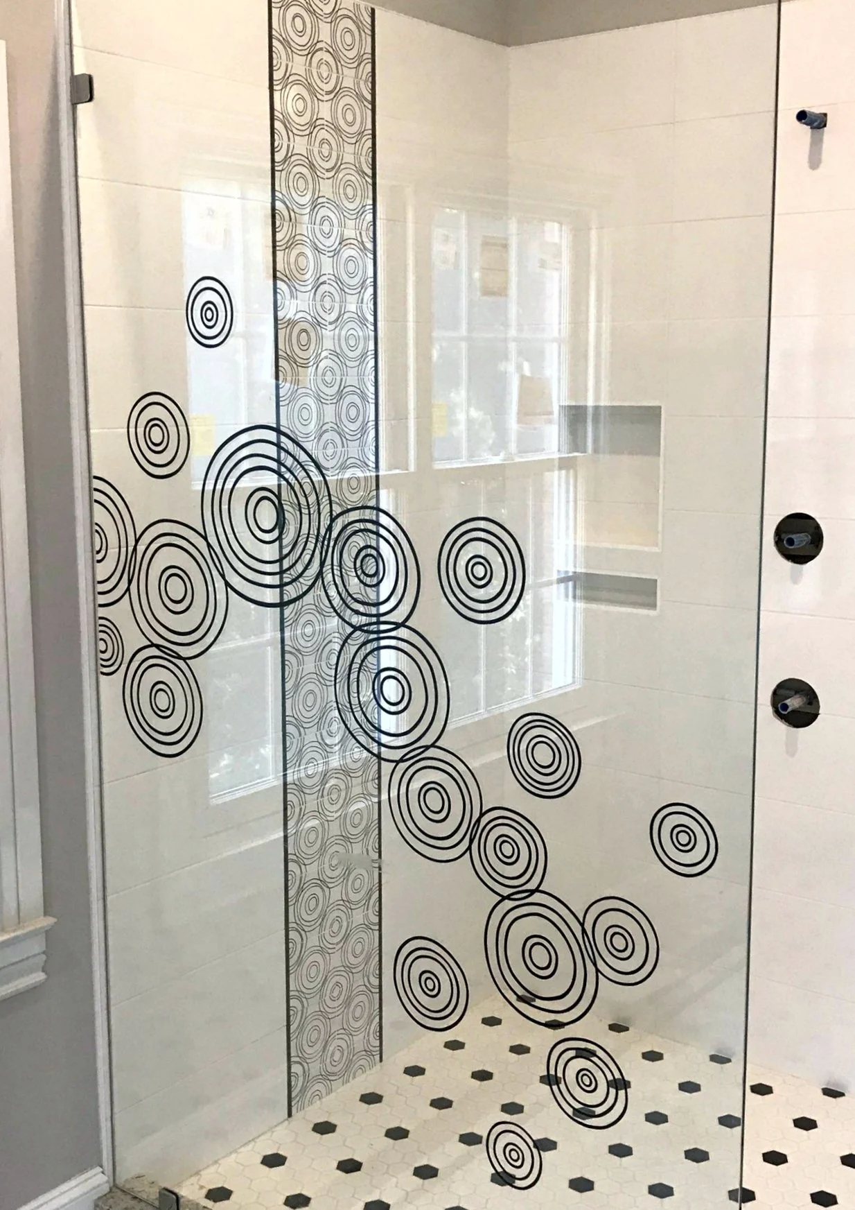 Glass shower door with black concentric circle patterns, white tiled walls, and a window reflecting outside scenery.