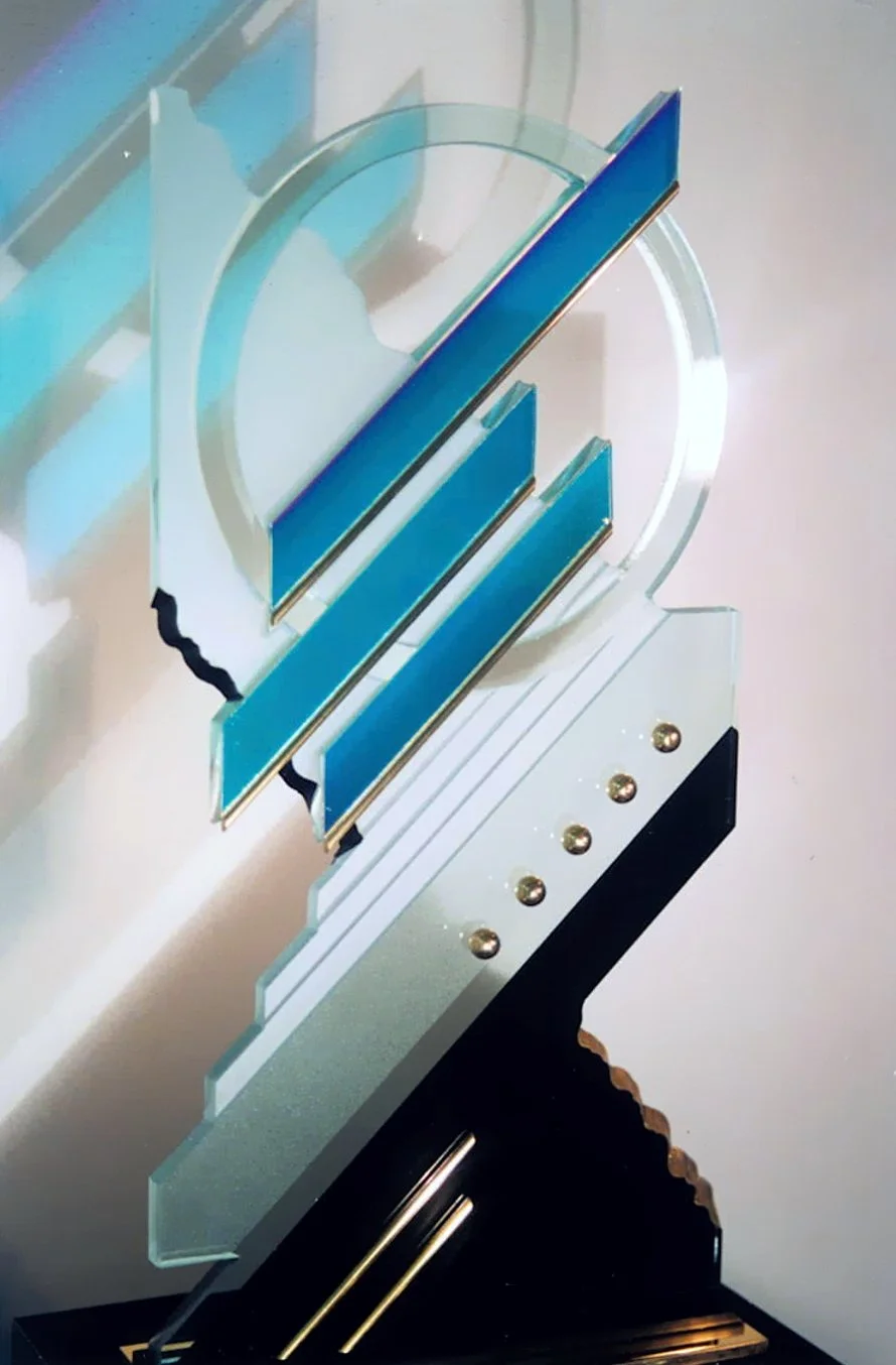 Close-up image of a glass and metal award or trophy with blue accents, featuring a sleek modern design with layered elements and gold screws, standing on a black base.