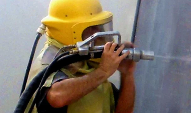 Construction worker in a yellow hard hat and protective gear operating a large drilling or cutting tool against a wall.