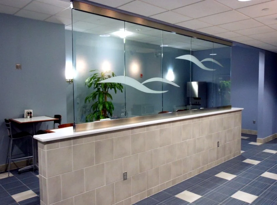 Interior view of a modern office area with an etched glass partition featuring a wave design, a plant, a small table with chairs, and tiled flooring.