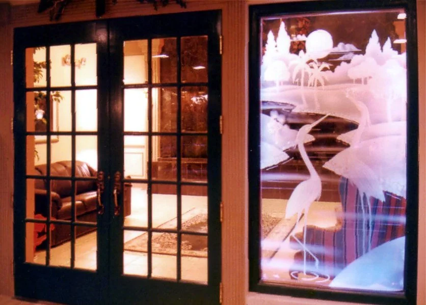 Double glass doors leading into a country club with leather sofas and a patterned rug; to the right, a large custom carved glass window with depicting a heron standing in water with trees and a moon, on a golf course background.