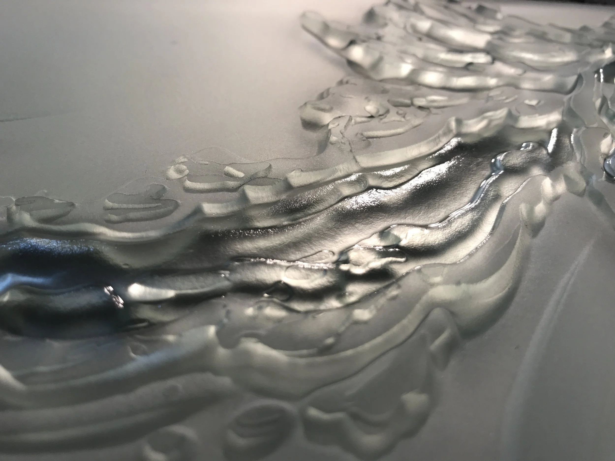 Close-up of metallic-silver, textured abstract artwork with layered, curving shapes on a smooth surface.