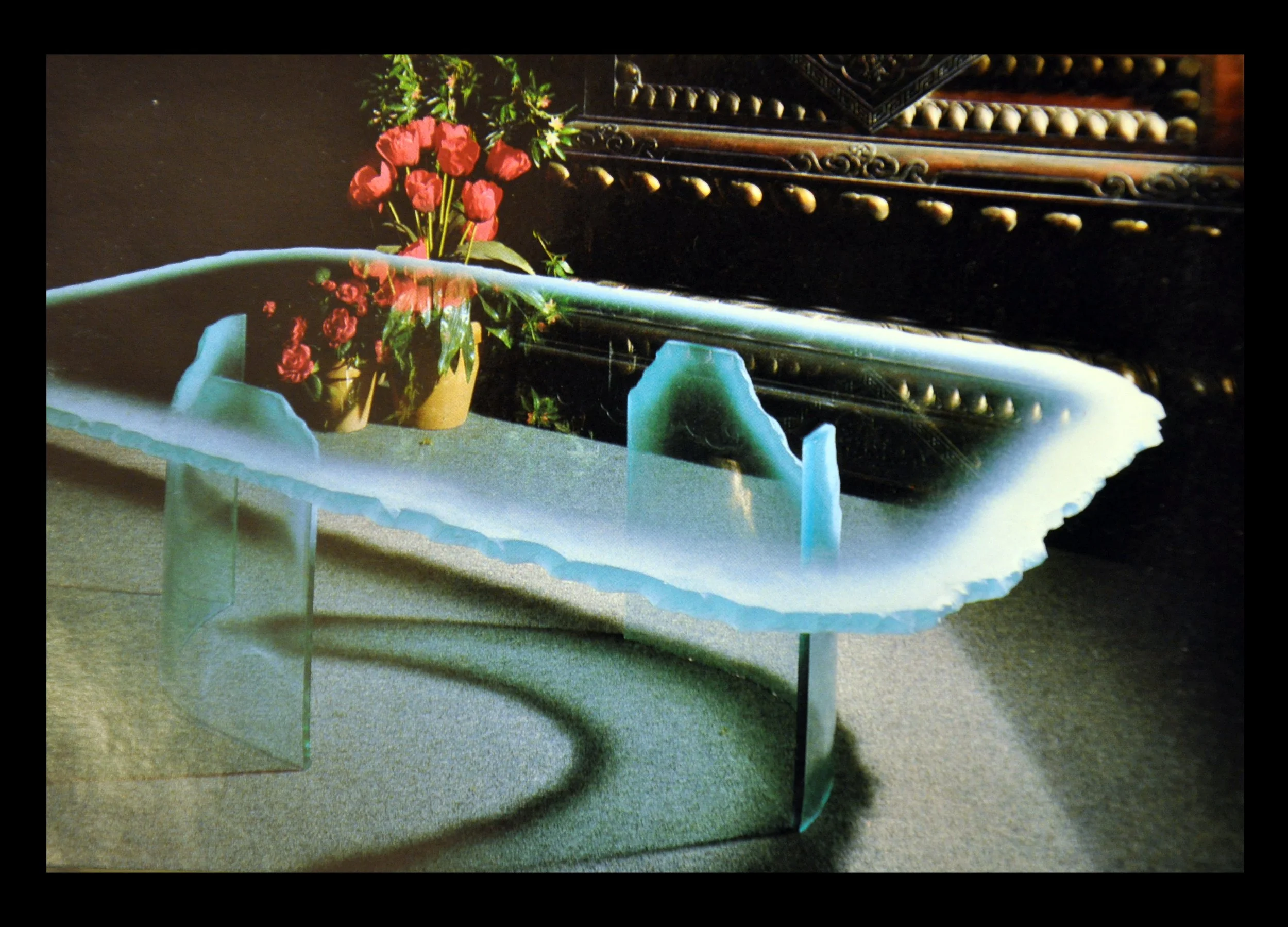A glass table with an irregular edge and a bent glass base, positioned on a carpeted floor. Behind the table, there is a potted plant with pink flowers and a dark, ornate wooden piece of furniture.