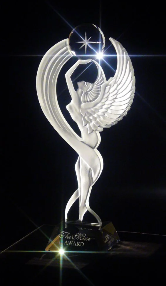 Clear crystal award sculpture with a winged woman holding a sphere with a star inside, illuminated with light reflections, and the base labeled 'The Muse Award'.
