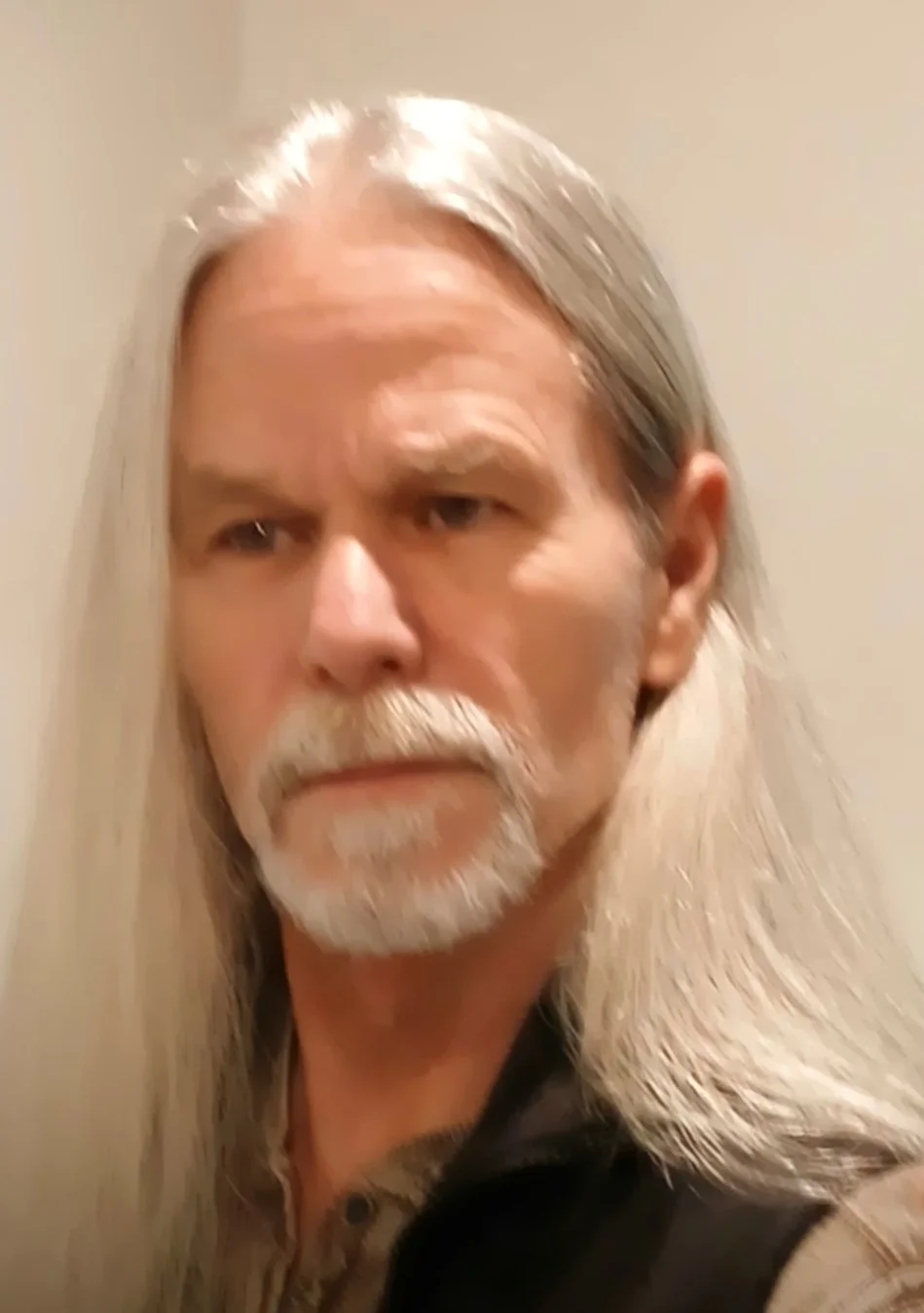 A man with long gray hair and a gray beard looking serious, standing against a plain beige wall.