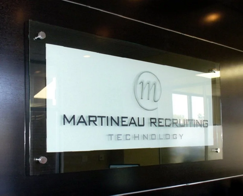 Layered glass sign with the logo for Martineau Recruiting Technology mounted on a dark wall.