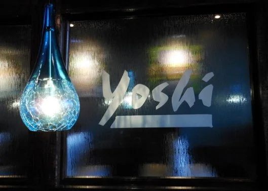 A sign with the word 'Yoshi' and a blue hanging light fixture glowing near a window.