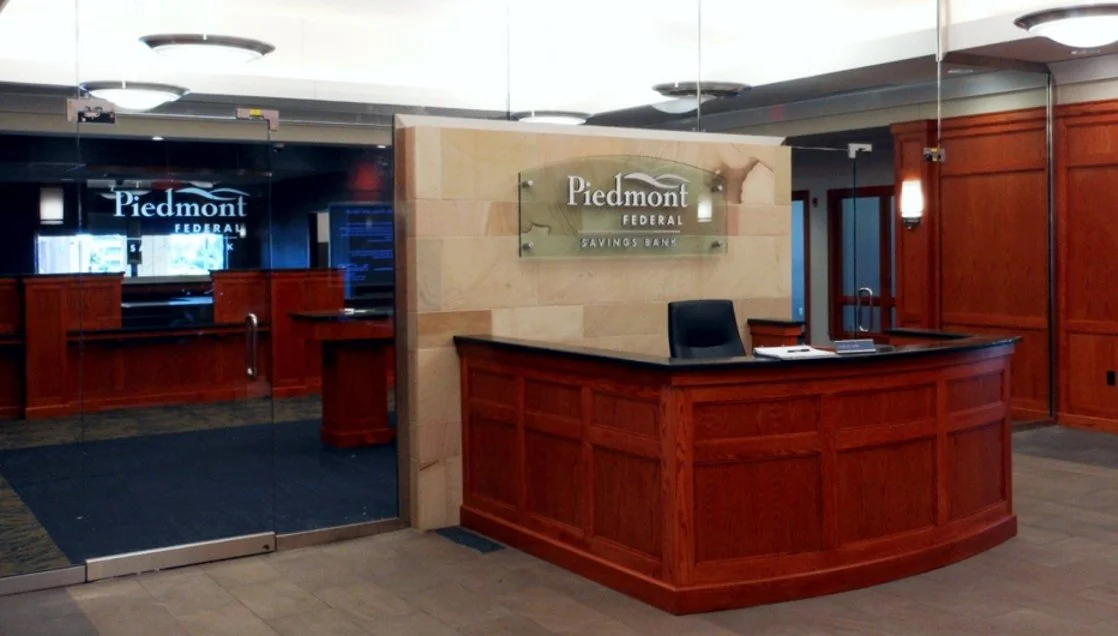 Inside the Piedmont Federal Savings Bank, featuring a wooden reception desk with a custom-made thick glass sign on a stone wall behind the desk, and wood-paneled walls.
