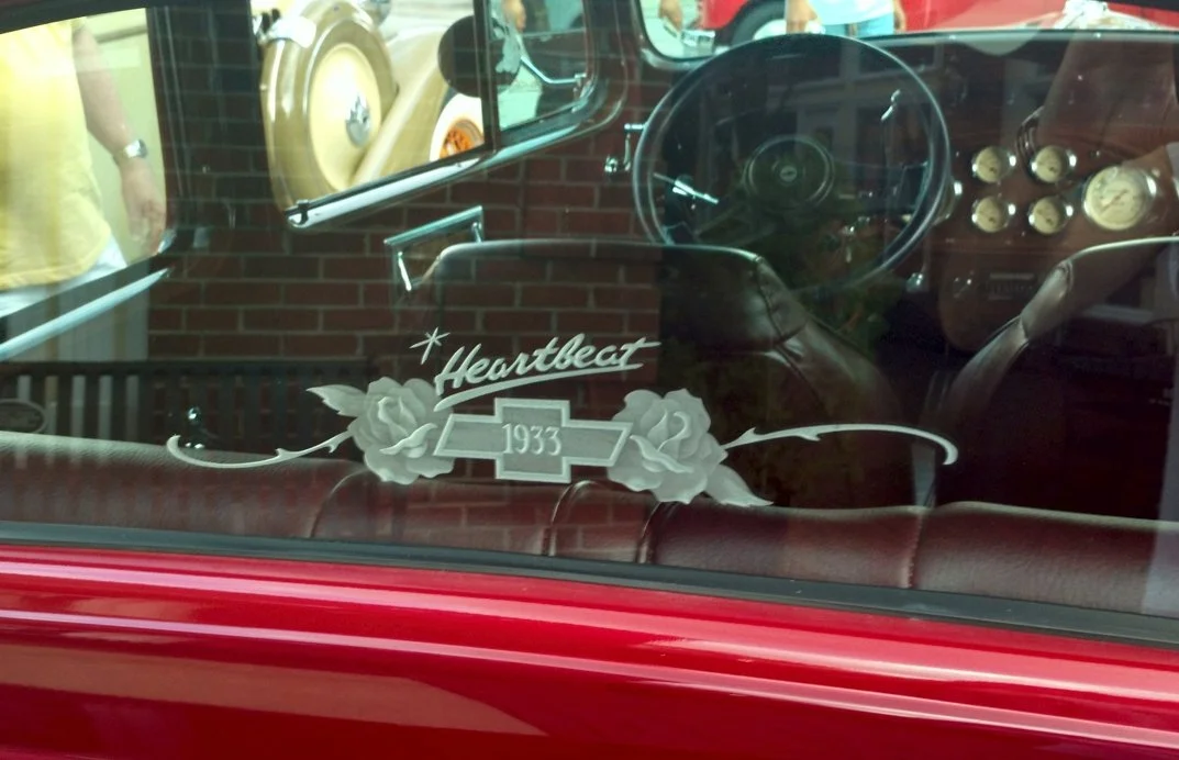 View of a vintage car window showing glass etching on the window that reads 'Heartbeat 1933' with roses and a Chevrolet logo.