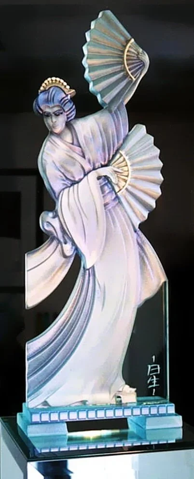 A detailed, stylized glass sculpture of an oriental woman with pearl finish, holding fans, dressed in flowing robes, set on a mirror pedestal with a glass base.