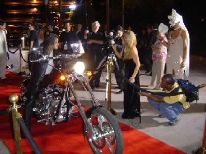 Reporters gathered at a red-carpet event with artist Michael Ray riding a motorcycle down the red carpet, photographers and others.