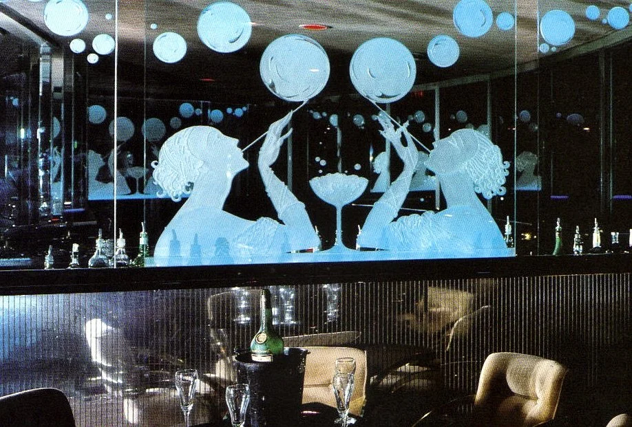 A mirror mural of two women with curly hair clinking martini glasses, surrounded by floating bubbles, in a bar setting with bottles and glassware.