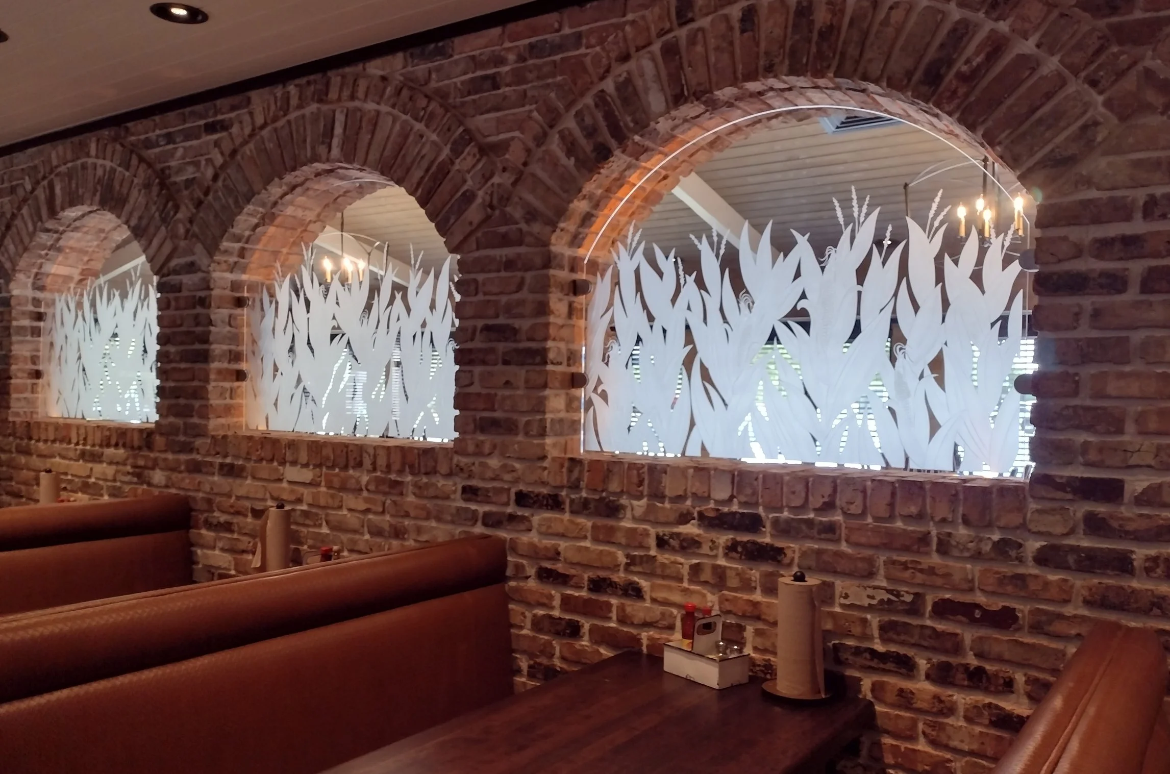 Interior of a restaurant with brick walls, arched openings with decorative carved glass panels featuring a corn crop design, wooden booths, and dining tables.