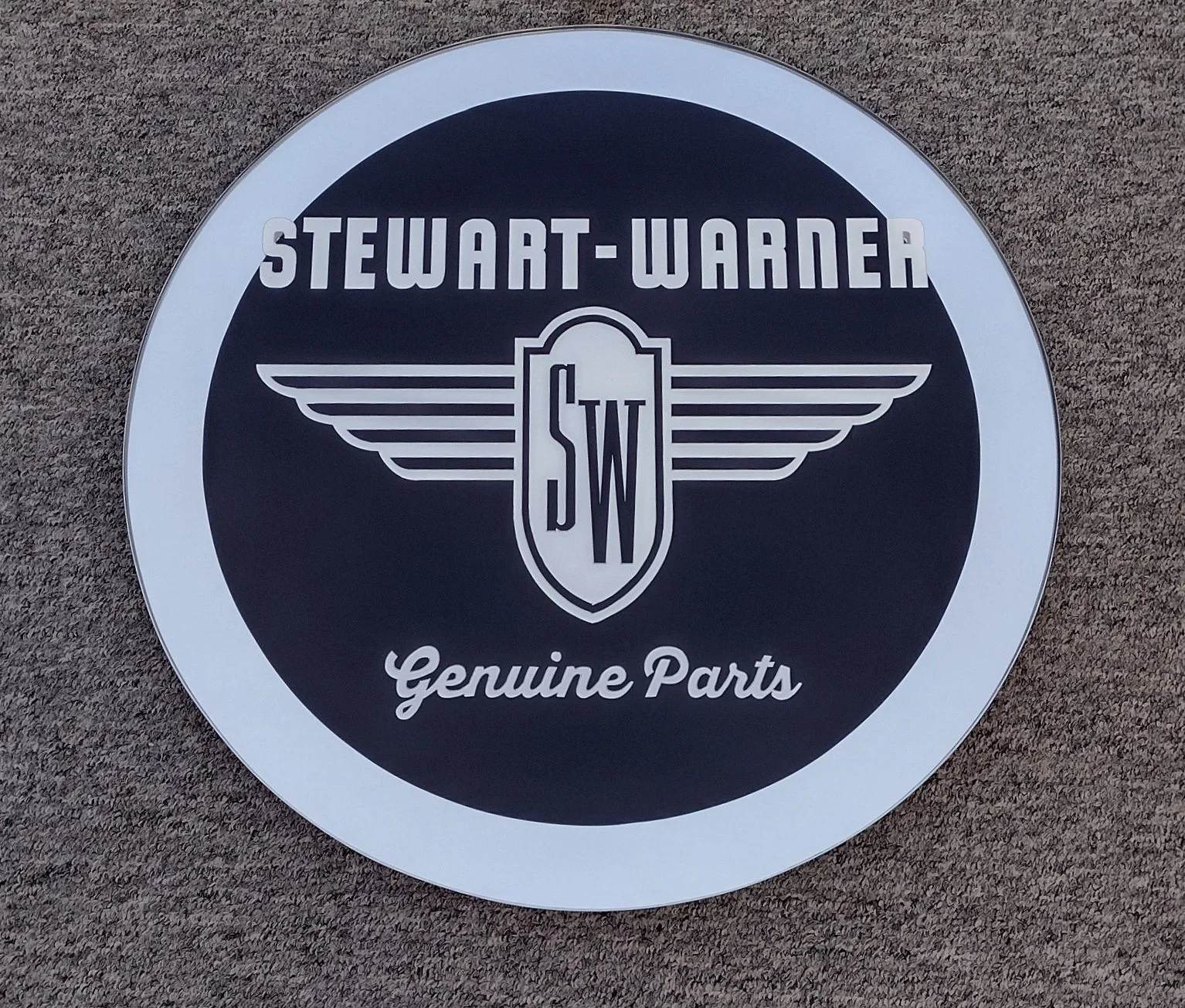 A circular sign with black background and white border, displaying the Stewart-Warner logo with wings, and the words 'Genuine Parts' at the bottom.