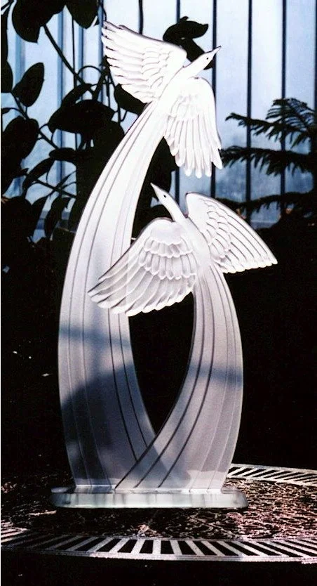 Decorative metal sculpture of two white flying swans with outstretched wings, set against a backdrop of greenery and a metal fence.