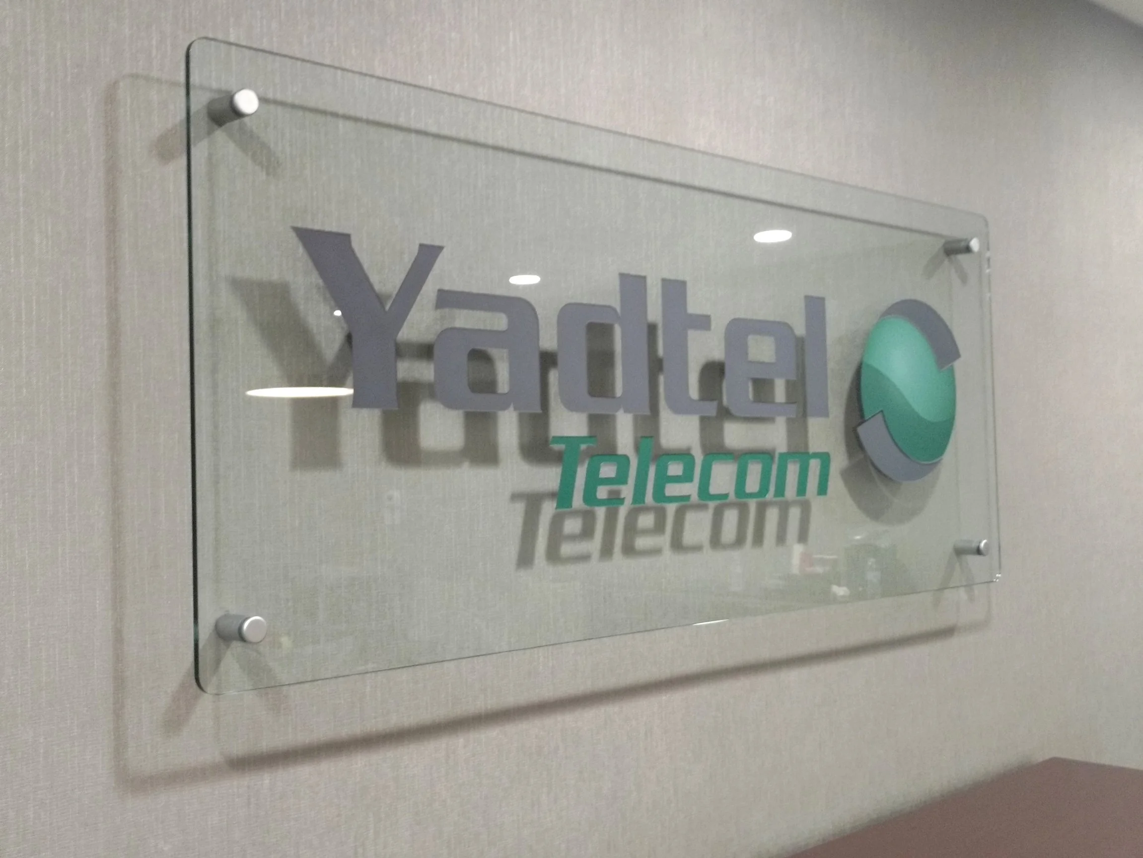 Yadtel Telecom logo carved and painted on a clear thick glass sign mounted on a beige wall.