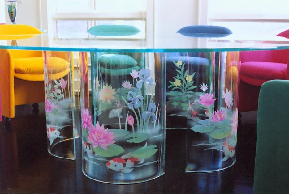 A large 96" diameter glass dining table with colorful chairs around it. The bent glass table bases have decorative carved and painted images of koi fish, water lilies, and lotus flowers.