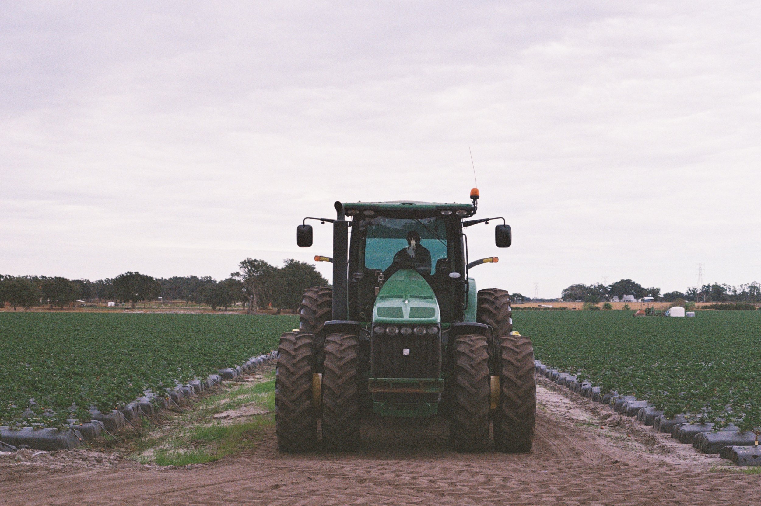 A green tractor with a person inside, on a farm with fields of crops and farm structures in the background, under a cloudy sky.