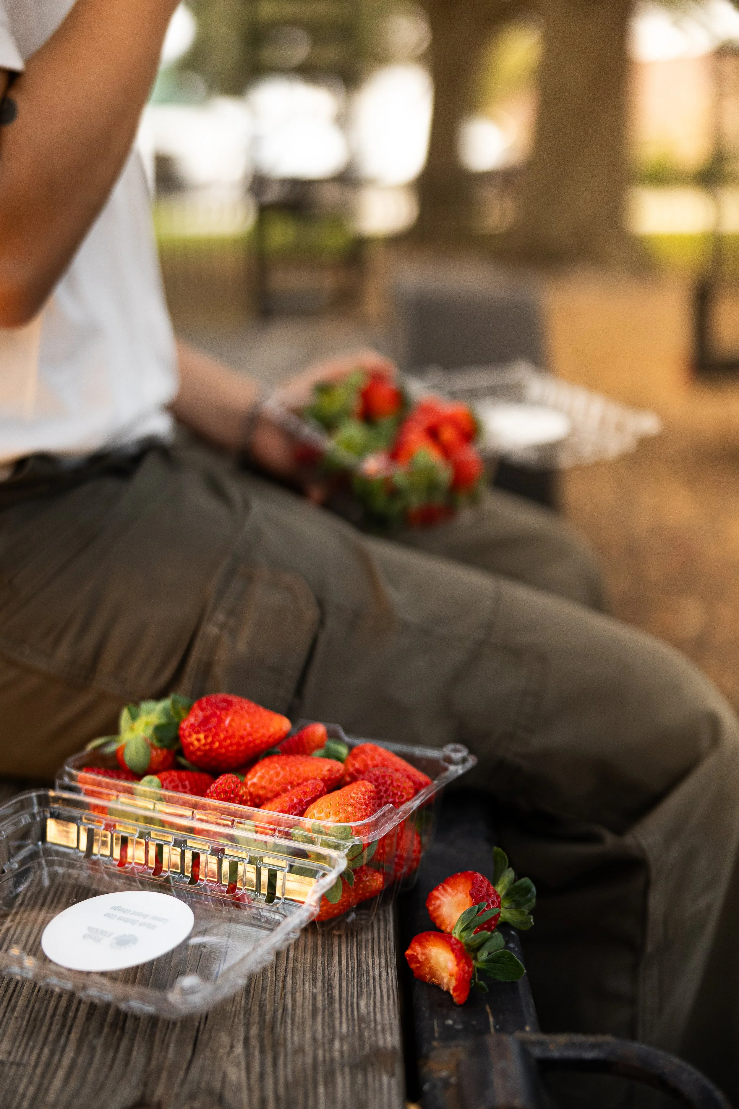 Close-up of a plastic container filled with fresh strawberries on a wooden table, with some strawberries and strawberry leaves beside it. In the background, a person is holding more strawberries, sitting outdoors with trees and a park-like setting.