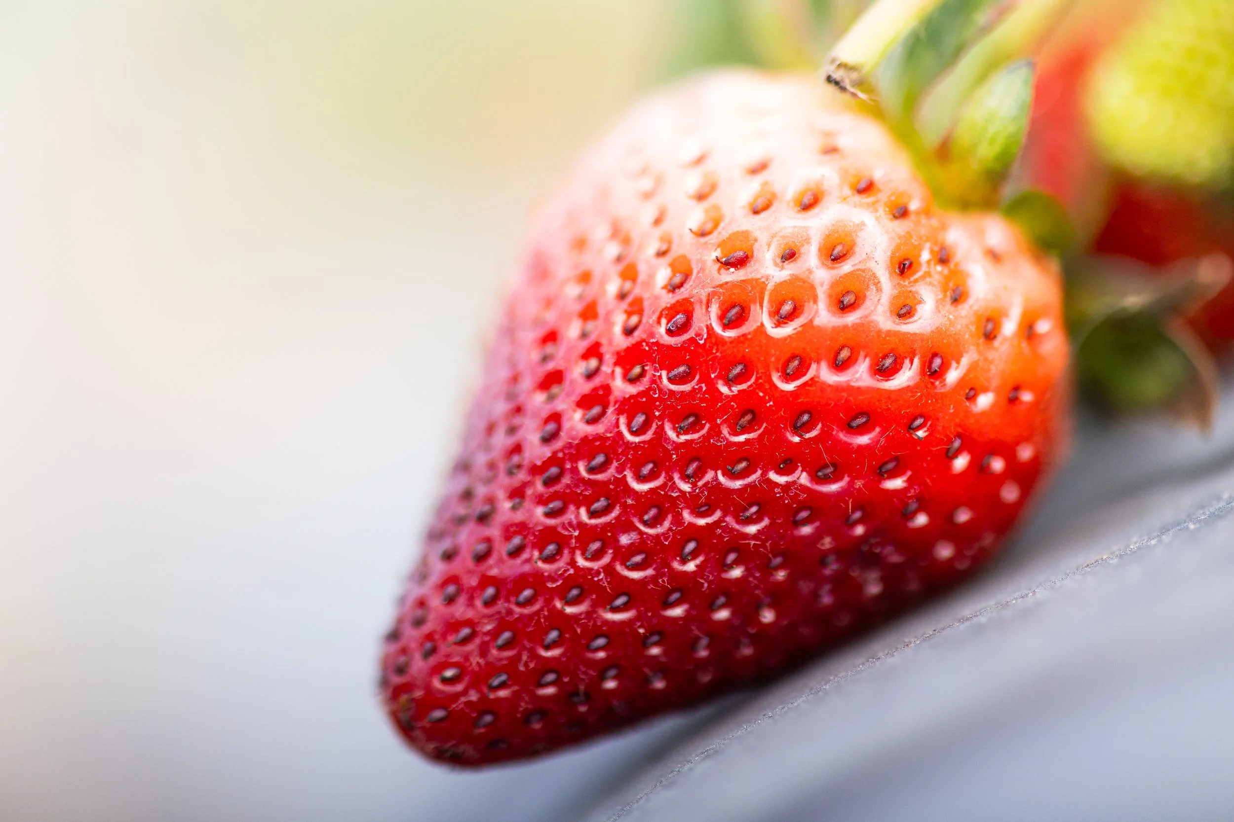 Close-up of a ripe, red strawberry with green leaves at the top, on a blurred light background.