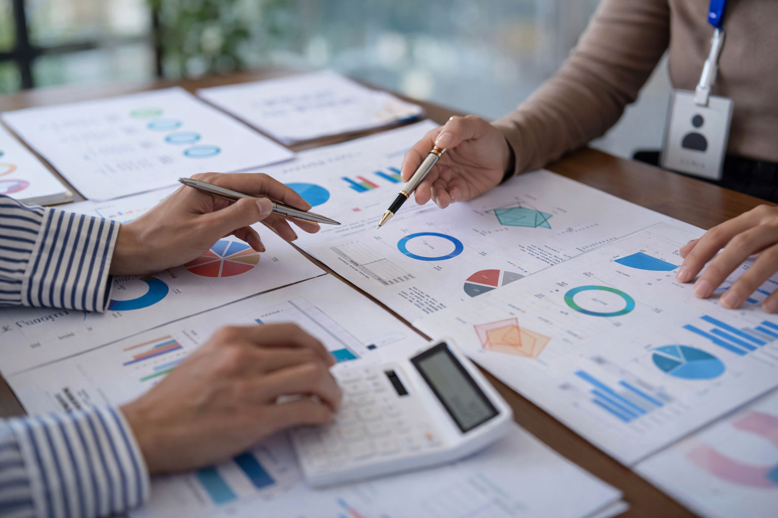 People discussing financial or business reports with various charts and graphs on papers, using pens and a calculator on a wooden table.