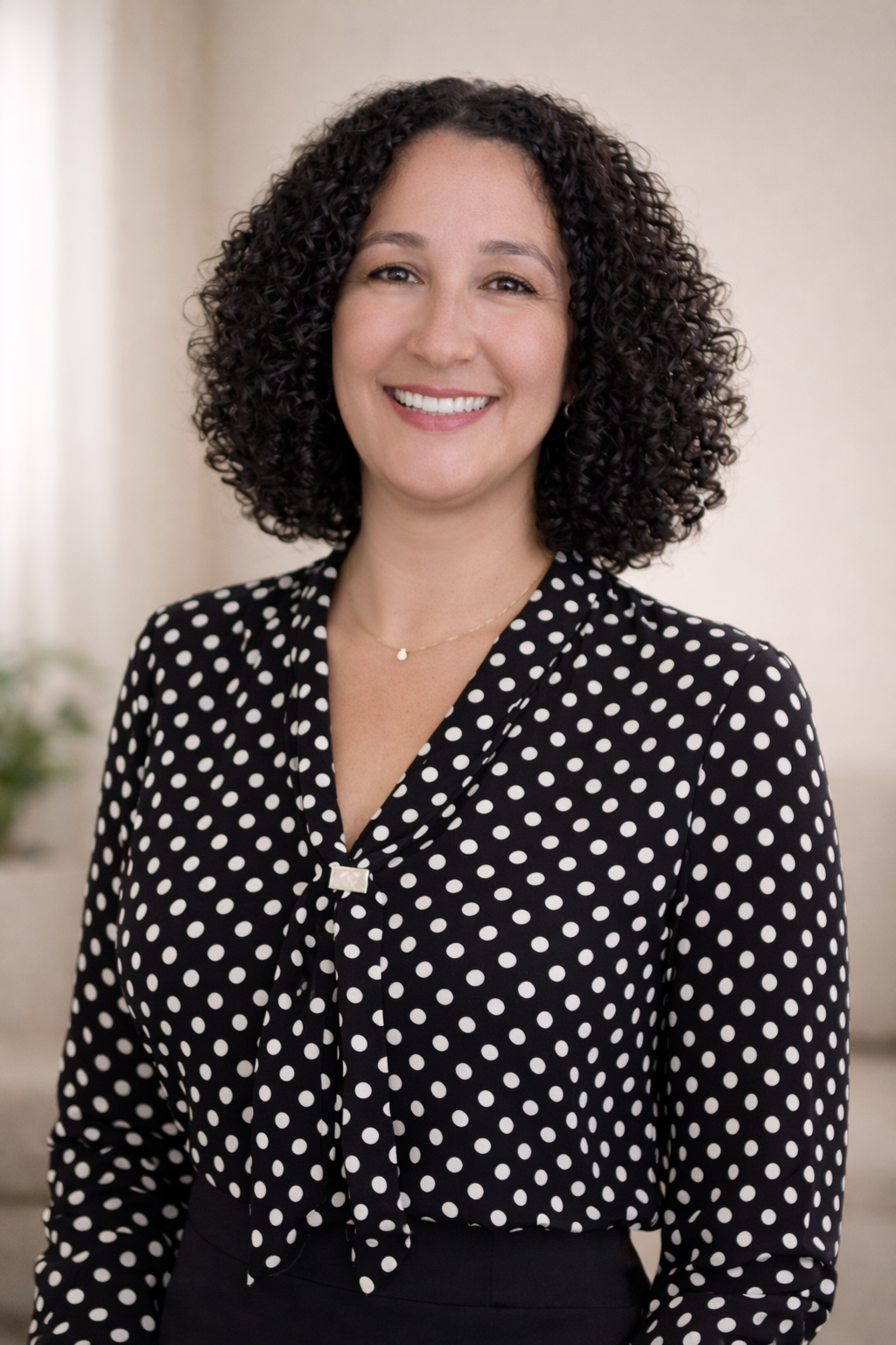 A woman with curly dark hair smiling, wearing a black and white polka dot blouse and a delicate necklace, standing indoors with a blurred background.