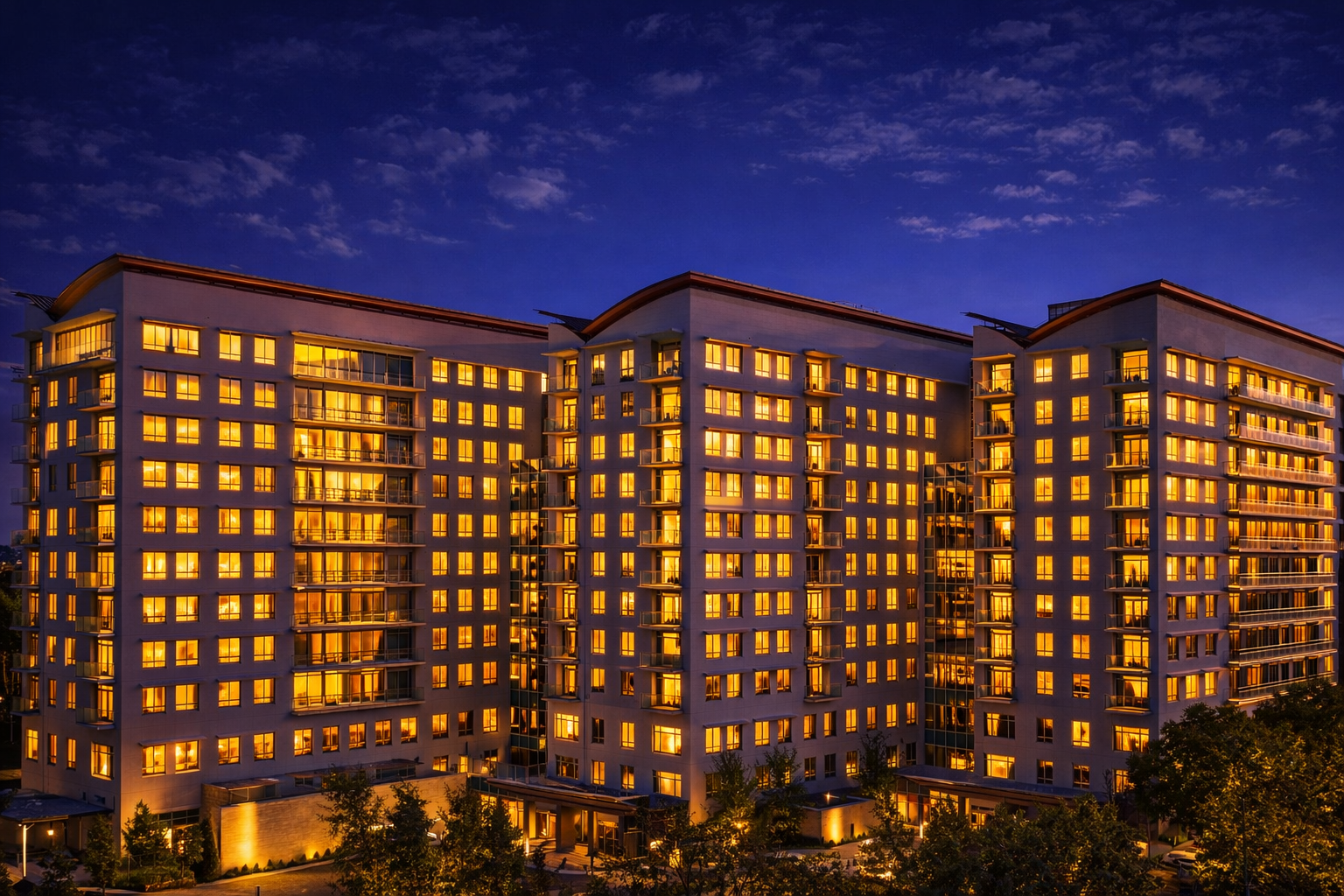 Modern multi-story apartment building at dusk with illuminated windows against a darkening sky.