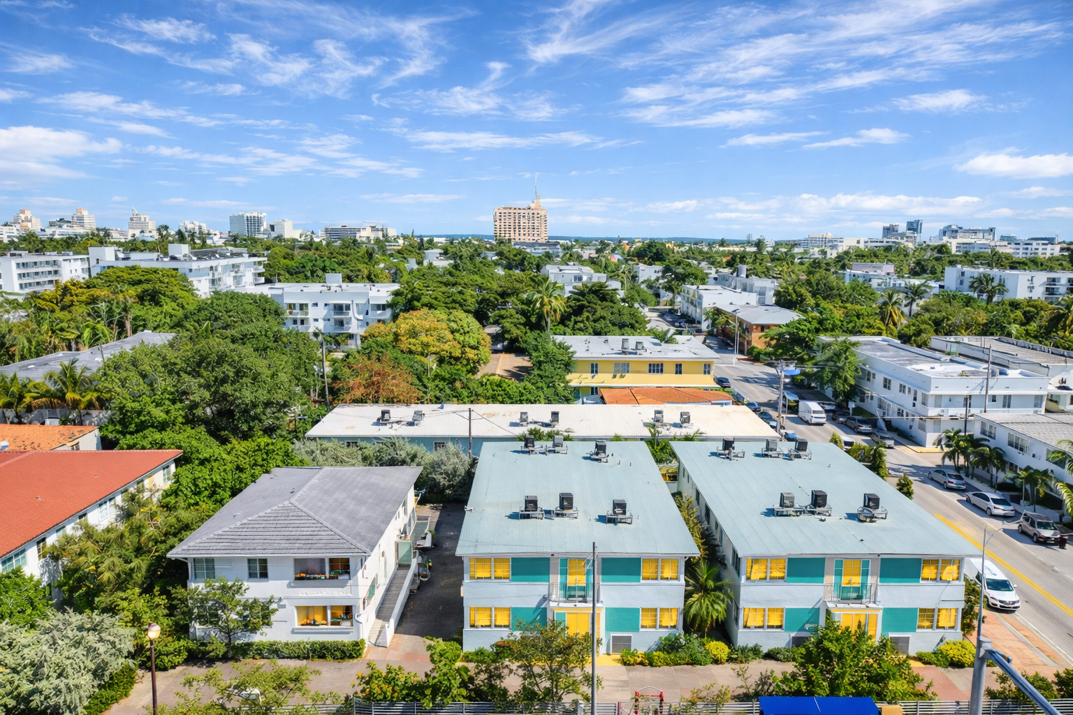 Aerial view of a neighborhood with colorful apartment buildings, surrounded by green trees and parked cars, under a partly cloudy blue sky.
