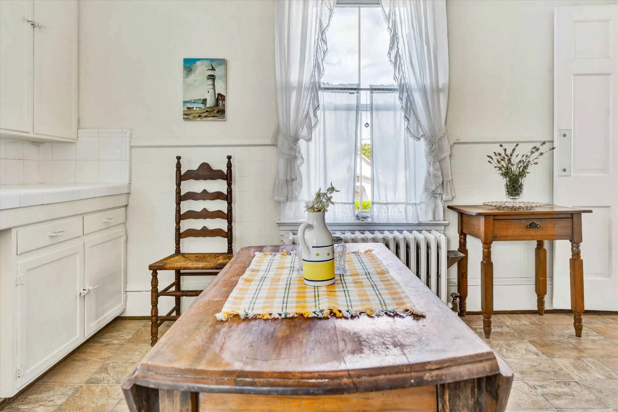 A cozy kitchen with a wooden dining table, a window with white curtains, and simple decorations including a painting of a lighthouse on the wall, and a vase with flowers on a side table.